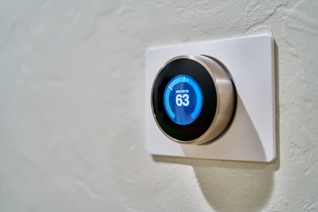 A photo of a digital smart thermostat on the wall. It is set to 63°F.